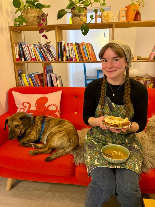 Junge Frau mit Zöpfen hält eine Schüssel Essen, während ein Hund auf der Couch liegt.
